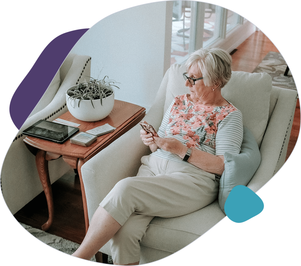 Elderly woman in an armchair using her smartphone, with a side table holding a tablet and a plant